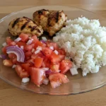 Chicken & Rice Meal with Tomato Salad on a white plate with fresh herbs and lemon wedges